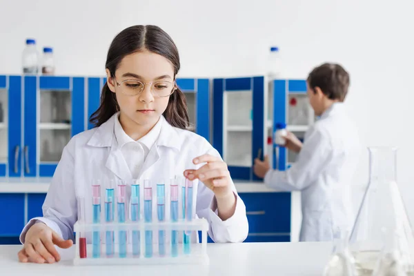 Fille en manteau blanc et lunettes près des éprouvettes avec liquide et garçon sur fond flou — Photo de stock