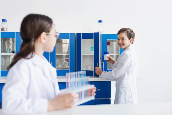 Sonriente chico en blanco abrigo celebración jar con polvo cerca chica con tubos de ensayo en borrosa primer plano - foto de stock