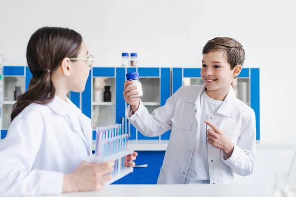 Glücklicher Junge zeigt auf Glas mit weißer Substanz neben verschwommenem Mädchen mit Reagenzgläsern — Stockfoto