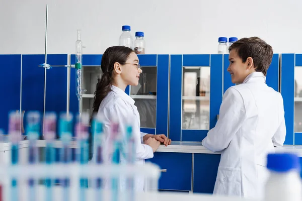Amigos sorridentes conversando perto do armário e tubos de ensaio em primeiro plano desfocado no laboratório — Fotografia de Stock