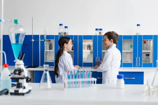 Side view of kids in white coats talking near test tubes and microscope on desk — Stock Photo