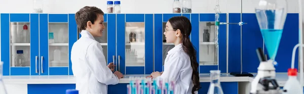 Вид сбоку детей в белых халатах, смотрящих друг на друга возле шкафчика в химической лаборатории, баннер — стоковое фото