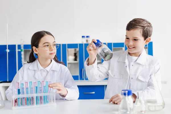 Alegre chico celebración tarro con sustancia química cerca chica en gafas y tubos de ensayo en laboratorio - foto de stock