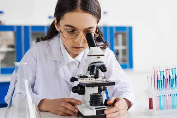 Brunette girl in white coat and eyeglasses using microscope in lab — Stock Photo