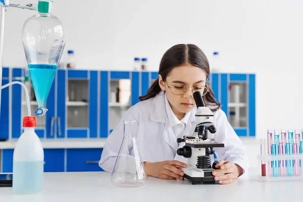 Preteen girl in eyeglasses looking in microscope near test tubes and flasks — Stock Photo