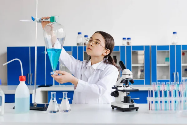 Preteen girl in eyeglasses and white coat holding flask near microscope and test tubes — Stock Photo