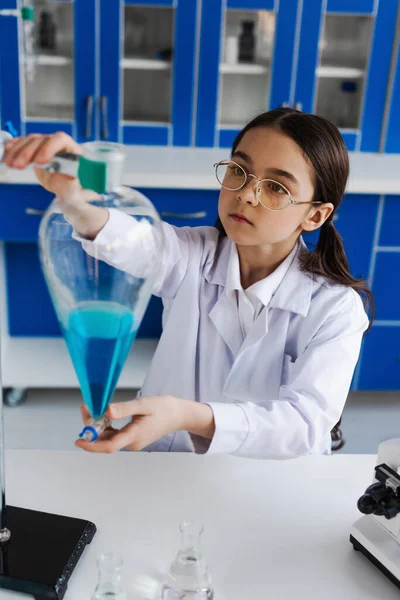 Mädchen mit Brille und weißem Kittel hält Kolben mit blauer Flüssigkeit im Labor — Stockfoto