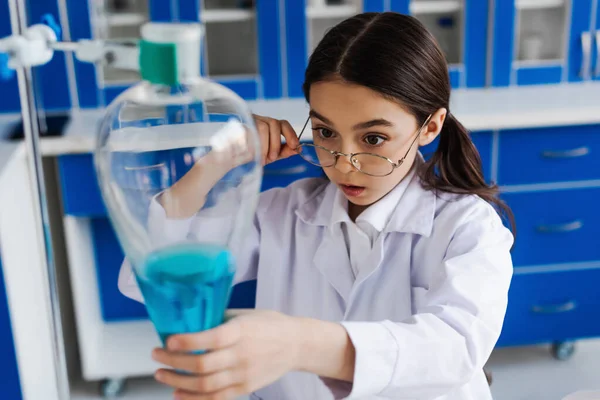 Preteen girl holding eyeglasses while looking at blurred flask with blue liquid during chemical experiment — Stock Photo