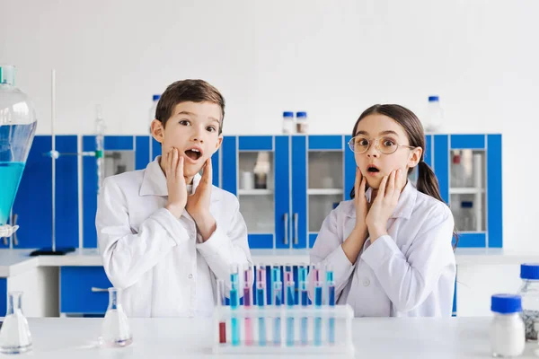 Schockierte Kinder in weißen Kitteln berühren Gesichter und schauen in Chemielabor in die Kamera — Stockfoto