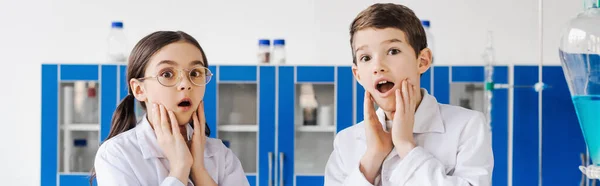 Bambini scioccati in camici bianchi che toccano i volti e guardano la fotocamera in laboratorio chimico, banner — Foto stock