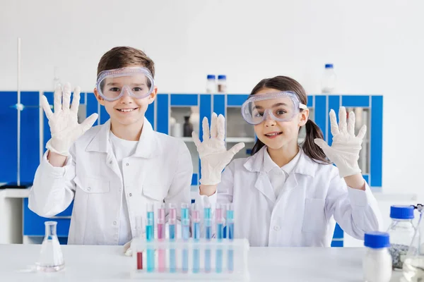Lächelnde Kinder in weißen Mänteln und Brillen, die Hände in Latexhandschuhen neben Reagenzgläsern im Labor zeigen — Stockfoto