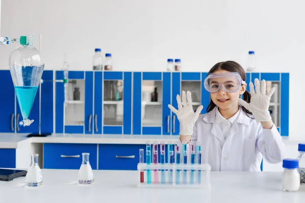 Lächelndes Mädchen mit Brille, das Hände in Latexhandschuhen in der Nähe von Kolben und Reagenzgläsern im Labor zeigt — Stockfoto
