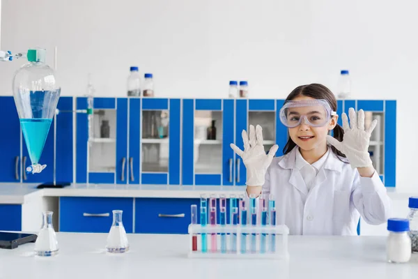 Smiling girl in goggles showing hands in latex gloves near flasks and test tubes in lab — Stock Photo