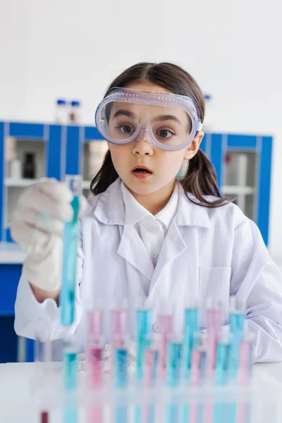 Stupito ragazza in camice bianco e occhiali guardando la provetta offuscata in laboratorio — Foto stock