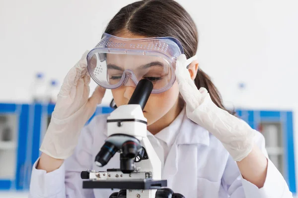 Preteen girl in latex gloves and goggles using microscope during chemical experiment — Stock Photo