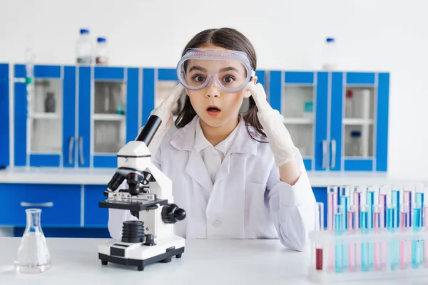 Amazed girl in goggles and latex gloves looking at camera near microscope and test tubes — Stock Photo