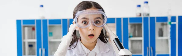 Shocked girl in goggles touching head and looking at camera in chemical laboratory, banner — Stock Photo