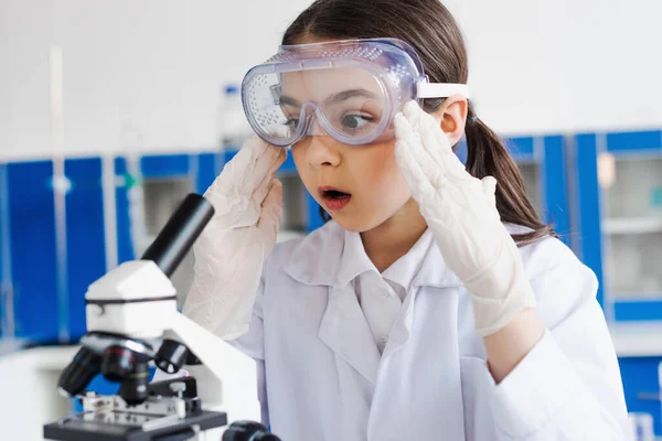 Schockiertes Mädchen mit Brille und Latexhandschuhen berührt Kopf nahe Mikroskop im Labor — Stockfoto