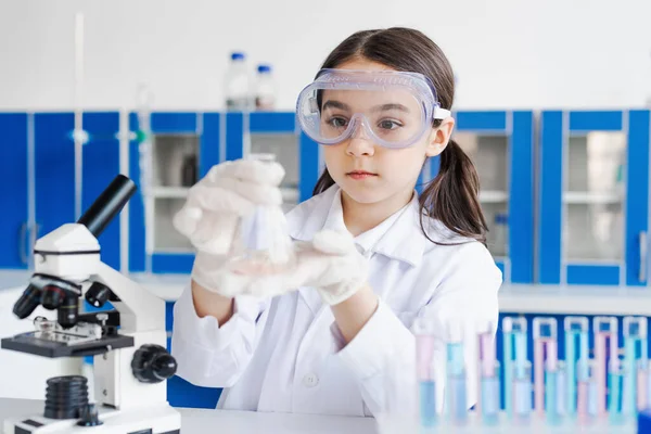 Niña con gafas sosteniendo frasco cerca del microscopio en el laboratorio - foto de stock