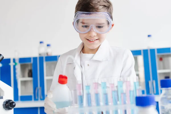 Niño sonriente con gafas sosteniendo la botella cerca de tubos de ensayo borrosos durante el experimento químico en el laboratorio - foto de stock