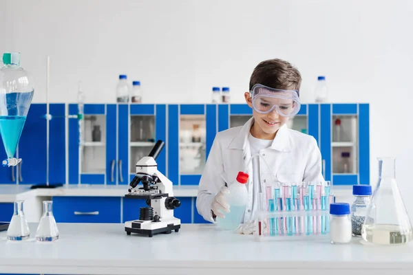 Niño feliz sosteniendo la botella cerca de tubos de ensayo y microscopio durante el experimento químico - foto de stock