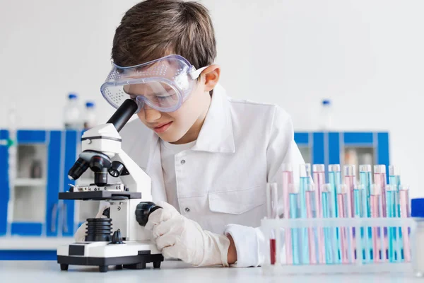 Preteen kid in goggles and latex gloves looking into microscope near test tubes — Stock Photo