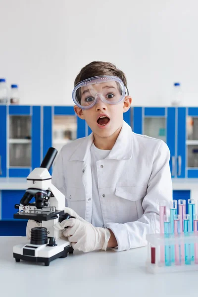 Amazed boy in goggles looking at camera near microscope and test tubes in laboratory — Stock Photo