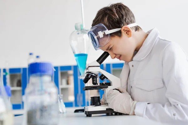 Side view of boy in white coat looking into microscope on blurred foreground — Stock Photo