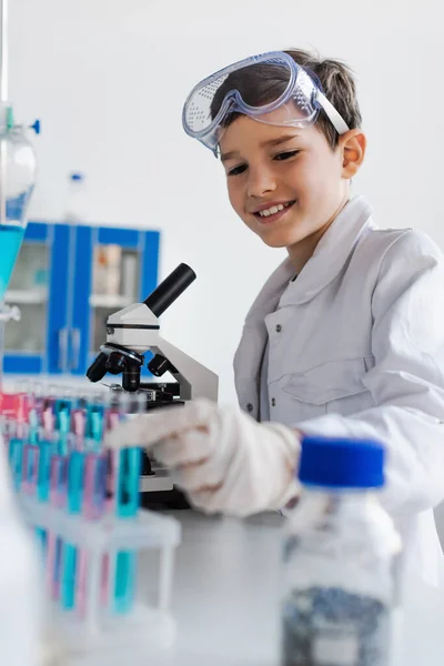 Cheerful boy with goggles smiling near blurred test tubes and microscope — Stock Photo