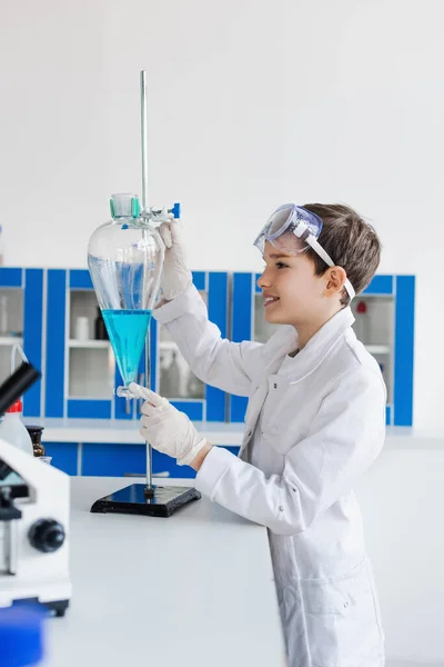 Side view of smiling kid in latex gloves looking at flask with blue liquid near microscope — Stock Photo
