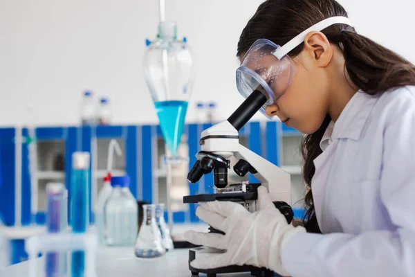 Side view of preteen girl in goggles looking into microscope in chemical laboratory — Stock Photo