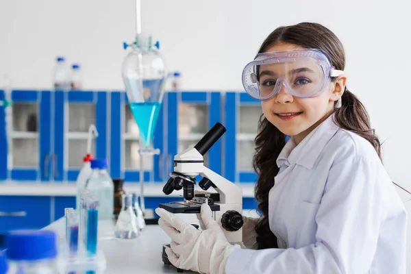 Preteen girl in goggles and white coat smiling at camera near microscope in lab — Stock Photo