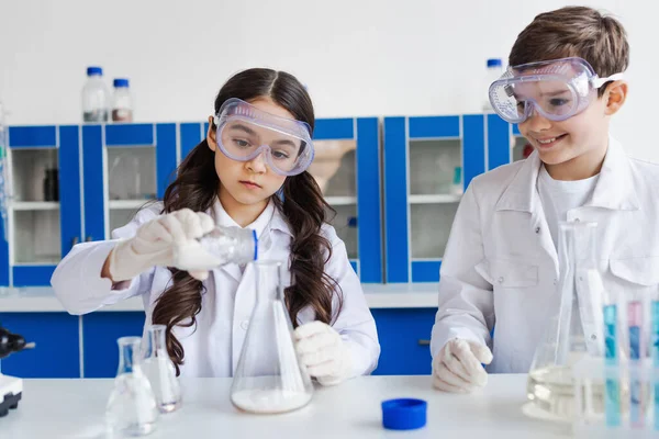 Chica seria en gafas vertiendo polvo en frasco cerca de un amigo sonriente en el laboratorio químico - foto de stock