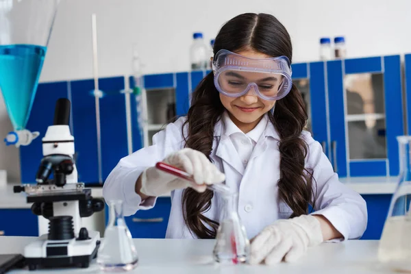 Happy brunette girl in goggles pouring red liquid from test tube into flask during chemical experiment in lab — Stock Photo