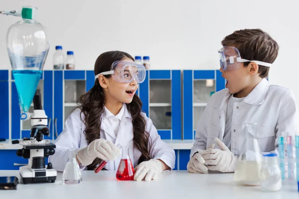 Amazed girl in goggles looking at friend near flask with red liquid in chemical lab — Stock Photo