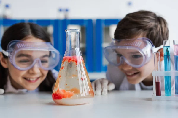Excited kids in goggles looking at flask with liquid and red substance in chemistry lab — Stock Photo