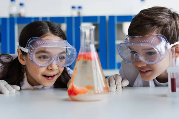 Здивовані діти в окулярах дивлячись на розмиту колбу з рідиною під час хімічного експерименту в лабораторії — стокове фото