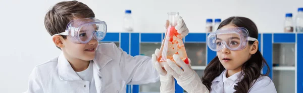 Ragazza sorpresa in maschera che tiene la fiaschetta con liquido vicino a un amico sorridente in laboratorio, banner — Foto stock