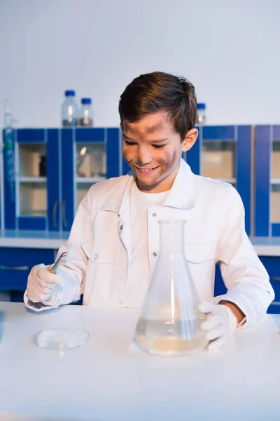 Niño sonriente con la cara sucia sosteniendo pinzas cerca de placa petri y frasco en el laboratorio - foto de stock