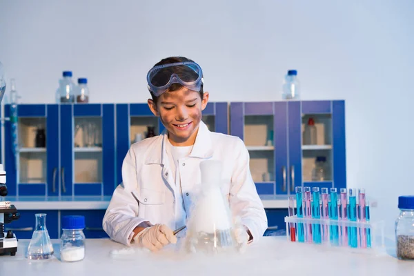Niño sonriente con la cara sucia mirando sustancias humeantes durante el experimento químico - foto de stock