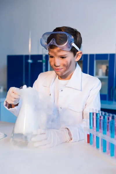 Niño preadolescente con la cara sucia sosteniendo matraz humeante mientras hace experimento químico en el laboratorio - foto de stock