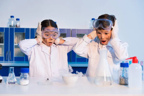 Bambini stupiti con facce sporche che toccano le teste mentre guardano ciotola fumante in laboratorio chimico — Foto stock