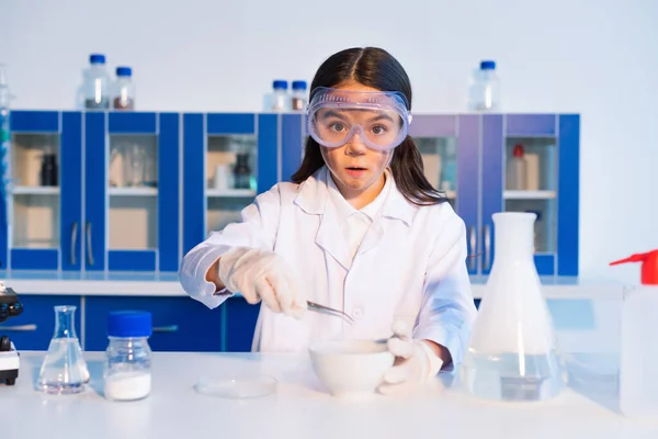 Surprised girl with dirty face holding tweezers near bowl and flask with steam in lab — Stock Photo