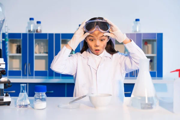 Chocó chica con la cara sucia sosteniendo gafas y mirando cuenco durante experimento químico - foto de stock