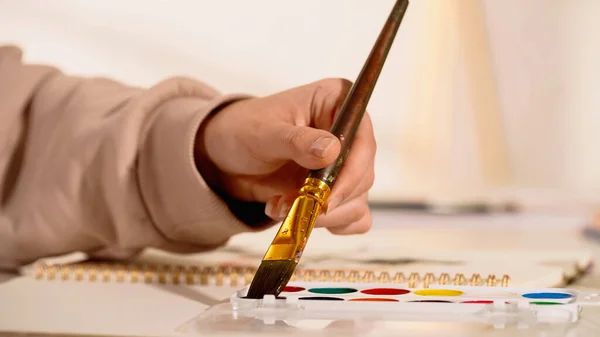 Vista recortada de la mujer sosteniendo pincel cerca de pintura acuarela y cuaderno de bocetos borroso - foto de stock