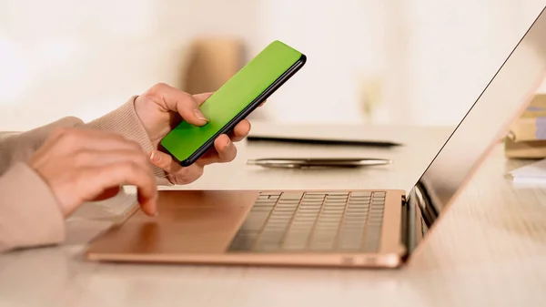 Vista recortada de freelancer usando smartphone e laptop com tela em branco em casa — Fotografia de Stock