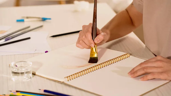 Cropped view of young woman painting on sketchbook near water and blurred color pencils on table — Stock Photo