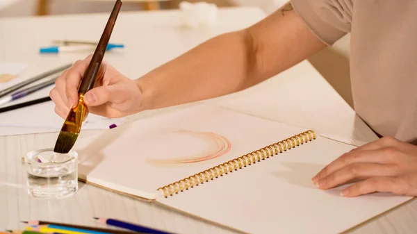 Vue recadrée d'une femme tenant un pinceau près d'un carnet de croquis et du papier sur une table — Photo de stock