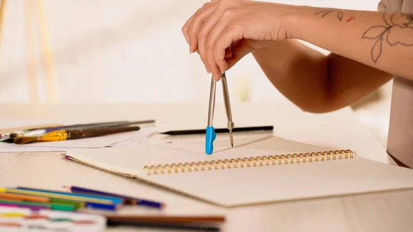 Vista cortada da mulher com tatuagem segurando a bússola de desenho perto do caderno de esboços em casa — Fotografia de Stock