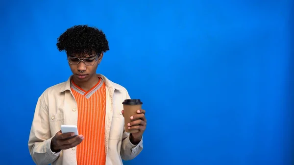 Giovane uomo afroamericano utilizzando smartphone e tenendo il caffè per andare su sfondo blu — Foto stock